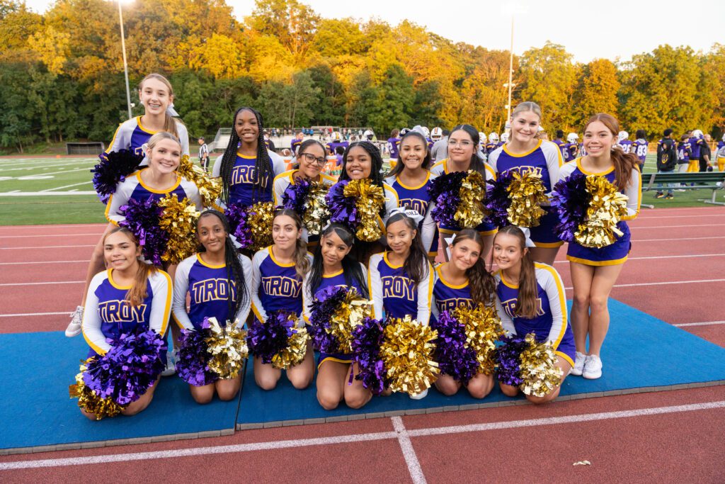 Sixteen Troy High School cheerleaders in purple and gold uniforms, posing with pom-poms on a track with a football field and trees behind them