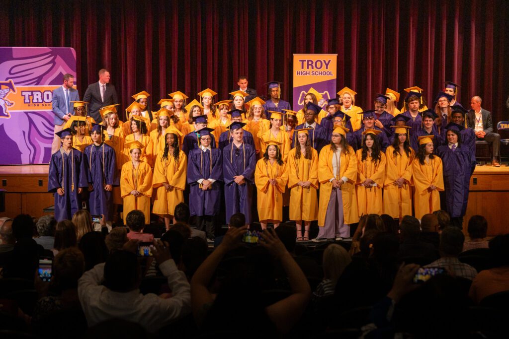 Graduating students from Troy High School in yellow and purple caps and gowns standing on stage during the ceremony, facing the audience.